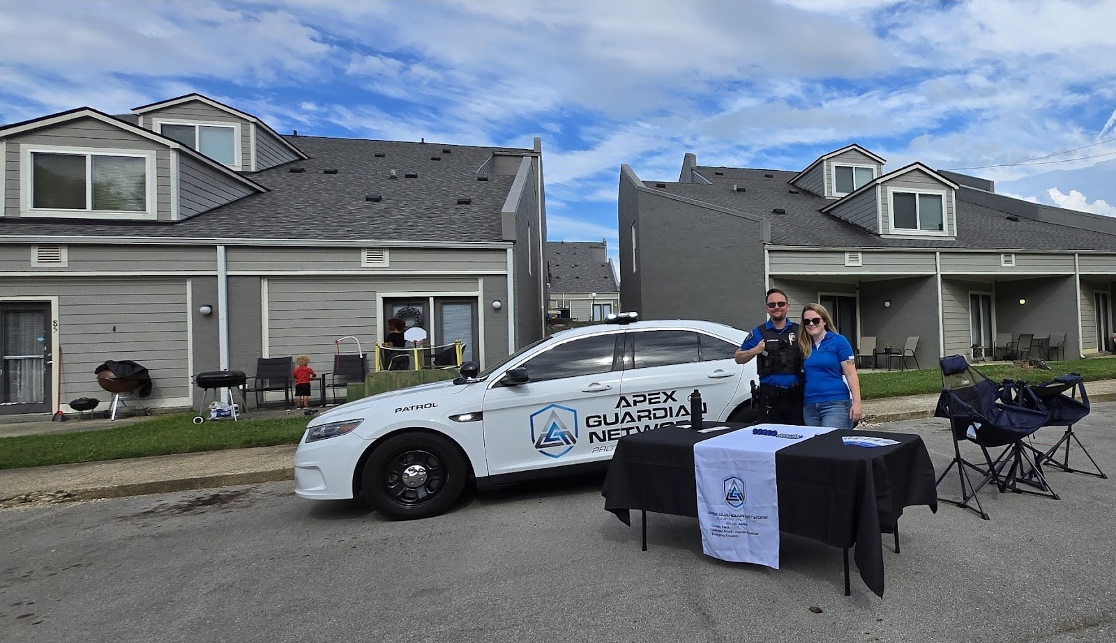 AGN officer and staff member at an outreach table beside the branded patrol car, talking with residents at an apartment community